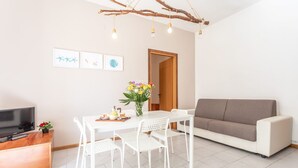 Una sala moderna con una mesa de comedor blanca, sillas blancas, un sofá marrón, un televisor sobre un mueble de madera y una lámpara con forma de rama.