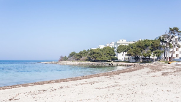 Una playa con arena, un mar tranquilo y una hilera de edificios blancos con árboles al fondo.