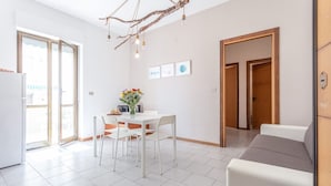 Un salón luminoso con una mesa blanca, sillas y un sofá. Hay una zona de comedor con una mesa y sillas, y una cocina con un refrigerador.