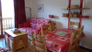 A cozy room with a red patterned sofa, a wooden dining table with chairs, a small desk with a chair, and a window with red curtains.