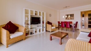 Una sala de estar con televisor, estanterías blancas, mobiliario de mimbre y una zona de comedor con sillas rojas.