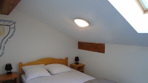 Une chambre avec un plafond en pente, un lit avec une literie blanche, une table de chevet avec une lampe et une petite étagère en bois.