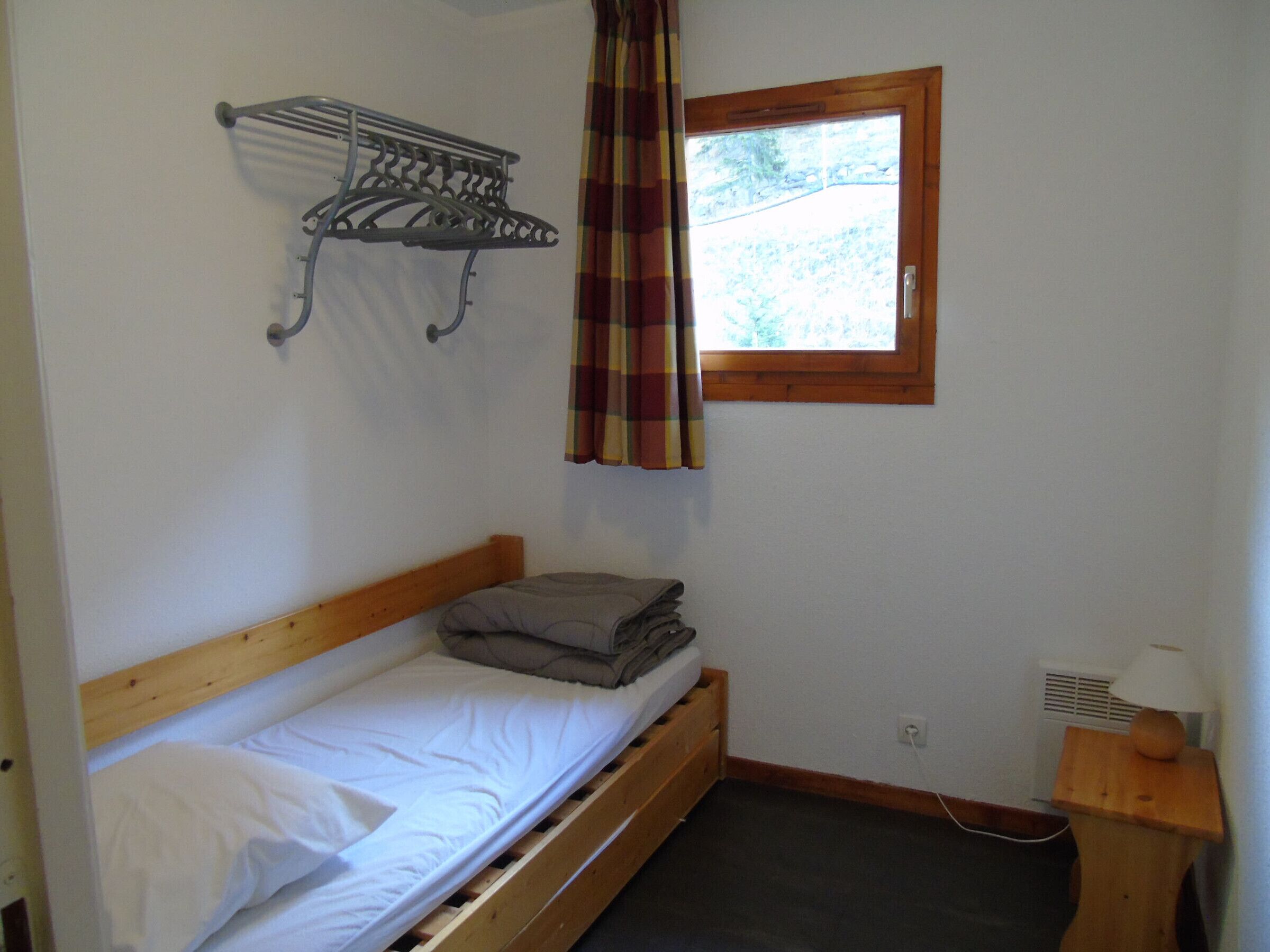 Une petite chambre avec un lit simple, une tête de lit en bois, un étendoir à linge en métal, une fenêtre avec un rideau à carreaux et une table de chevet avec une lampe.
