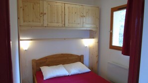 A small bedroom with a wooden bed frame, two white pillows, a red bedspread, and a wooden wardrobe above the bed.