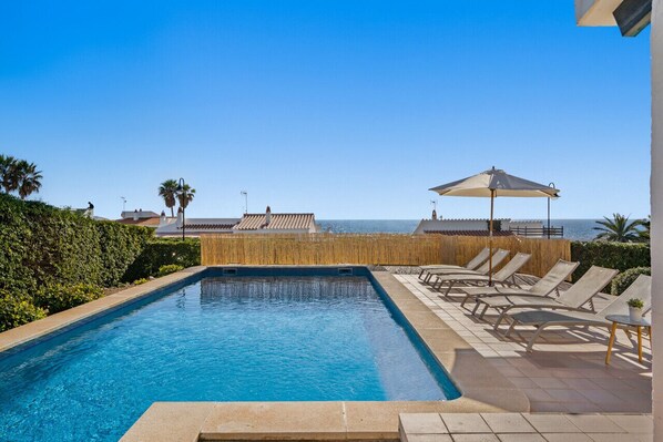 A swimming pool with lounge chairs and umbrellas, overlooking a coastal area with buildings and palm trees.