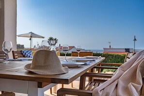 Une table en bois, dressée pour le dîner, avec un chapeau tressé, des verres à vin et une vue sur la mer.