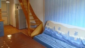 Ein Zimmer mit einer Holztreppe, einem Bett mit gemustertem Kopfteil, einer Küchenzeile mit Kühlschrank und einem blauen Vorhang.