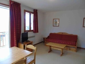 A small living area with a wooden table, a red sofa, a TV, and a window with curtains.