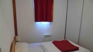 A small, simple room with a bed, a window with red curtains, and a wall-mounted heater. - Valfréjus: 2-room apartment, near ski lift, pet-friendly (Valfréjus)