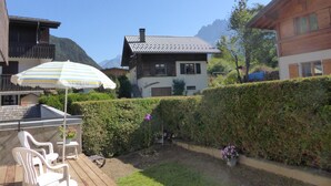 Une terrasse en bois avec des chaises en plastique blanches, un parasol à rayures et un jardin agrémenté d’une haie et de fleurs.