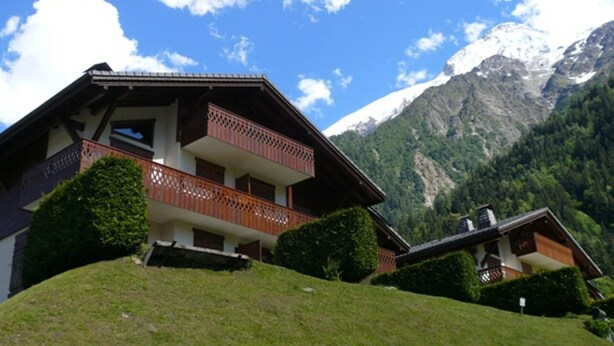 Un bâtiment de style chalet doté d’un balcon, entouré d’une végétation luxuriante et avec des montagnes en toile de fond.