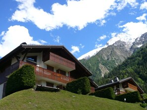 Edificio de estilo chalet con balcón, rodeado de exuberante vegetación y montañas al fondo.