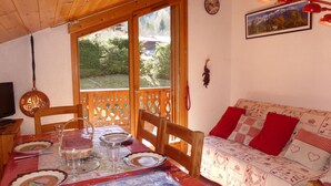 Une pièce chaleureuse avec une table à manger en bois, dressée pour quatre personnes, un canapé avec des coussins rouges, une horloge murale et un tableau encadré accroché au mur.