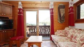 A wooden cabin interior with a TV, a sofa, a table, and a balcony with a view of the mountains. - 4-room apartment, pool and parking, near lifts (Les Houches)