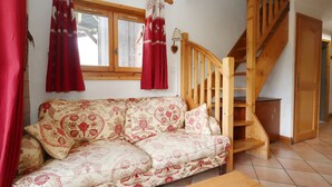 A cozy living room with a patterned sofa, a wooden staircase, and a window with curtains. - 4-room apartment, pool and parking, near lifts (Les Houches)