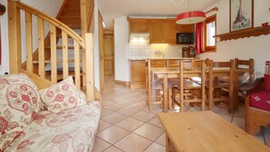 A cozy living room with a wooden staircase, a dining area with wooden furniture, and a kitchen with wooden cabinets and a red hanging light fixture. - 4-room apartment, pool and parking, near lifts (Les Houches)