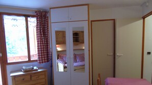 Une chambre avec une commode en bois, un lit recouvert d’une couverture rose, une armoire aux portes miroir et une fenêtre ornée de rideaux à carreaux.
