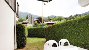 A patio with white plastic chairs and a table, surrounded by neatly trimmed hedges and a view of houses in the background.