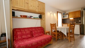 A cozy living room with a red sofa, wooden dining table, and chairs. There is a kitchen area with wooden cabinets and a microwave.