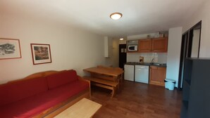 A compact living space with a red sofa, wooden table, and bench, a kitchenette with a microwave and dishwasher, and framed pictures on the wall. - Cozy 2-Room Apartment - Ski-In/Ski-Out, Balcony, Pet-Friendly, Sleeps 6 (Valfréjus)