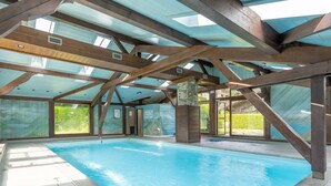 Una piscina interior con techo alto y vigas de madera, rodeada de amplios ventanales.