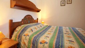 Un dormitorio con cabecera de madera, colcha con estampado floral, lámpara de noche y dos cuadros en la pared.