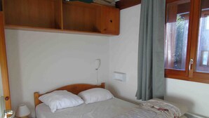 A small, single-bed room with a wooden headboard, a bedside table with a lamp, a small storage box, and a window with curtains. - Cozy apartment, pets welcome - 6 guests (Valfréjus)