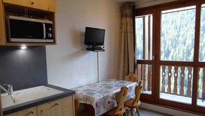 A small kitchen with a microwave, a sink, and a table with chairs. There is a TV on a stand and a window with a view of trees. - Cozy Studio for 4 near Ski Lifts and Shops, Includes Linens, Kitchen, Balcony with Scenic Views (Valfréjus)