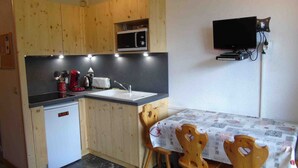 A compact kitchen with wooden cabinets, a microwave, a toaster, and a sink. There is a dining table with chairs and a television mounted on the wall. - Cozy Studio for 4 near Ski Lifts and Shops, Includes Linens, Kitchen, Balcony with Scenic Views (Valfréjus)