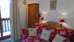 A cozy room with a red patterned sofa, a wooden cabinet, a wall clock, and two red lamps. - Cozy Studio for 4 near Ski Lifts and Shops, Includes Linens, Kitchen, Balcony with Scenic Views (Valfréjus)