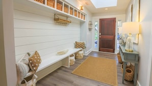 A hallway with a white bench, wicker baskets on shelves, a wooden door, and a lamp on a table.