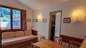Ein gemütliches Zimmer mit einer Holzsofa, einem kleinen Tisch und Stühlen. An der Wand hängen gerahmte Bilder und ein Fenster mit Vorhängen.
