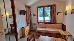 Ein gemütlicher Raum mit Holztisch und -stühlen, einer Schlafcouch, einem an der Wand montierten Fernseher und einem fenster mit Vorhängen.