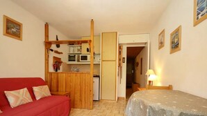 A compact room with a red sofa, a bed with a snowflake-patterned cover, a kitchenette with a microwave, and a wooden shelf.