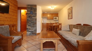 A cozy living room with a stone pillar, wooden furniture, a tiled floor, and a kitchen in the background. - 2-room apartment, 5 pers., skiing, parking, pool (Les Houches)