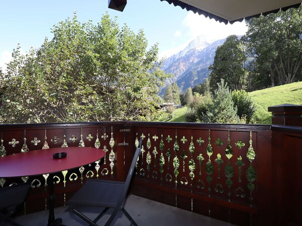 Een balkon met een rode tafel en stoelen, een decoratieve metalen hek en een bergachtig landschap op de achtergrond.