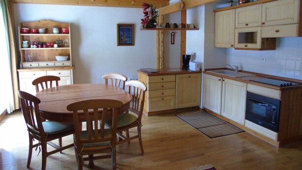 A wooden dining table with chairs, a kitchen with cabinets and appliances, and a clock on the wall.