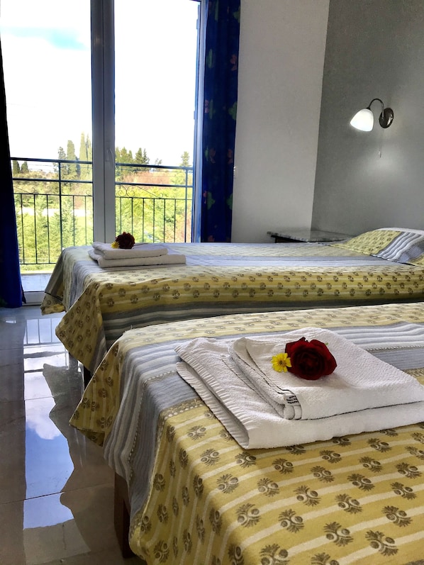 Habitación de hotel con dos camas, cada una con una colcha de motivos florales y una toalla blanca con una rosa roja. Se ve un balcón con barandilla metálica y vegetación a través de la ventana.