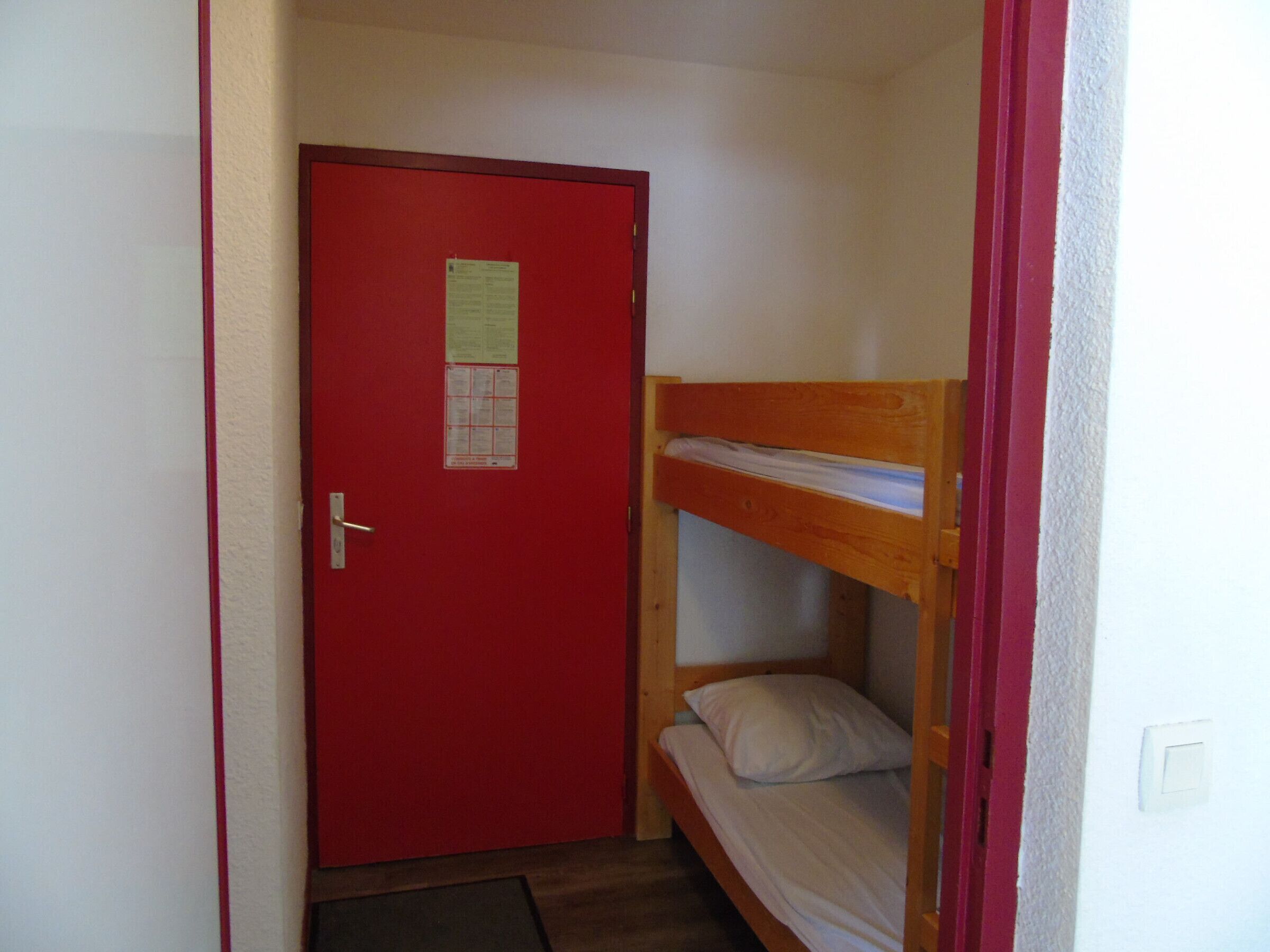 A small room with a red door, a bunk bed, and a small mat on the floor.