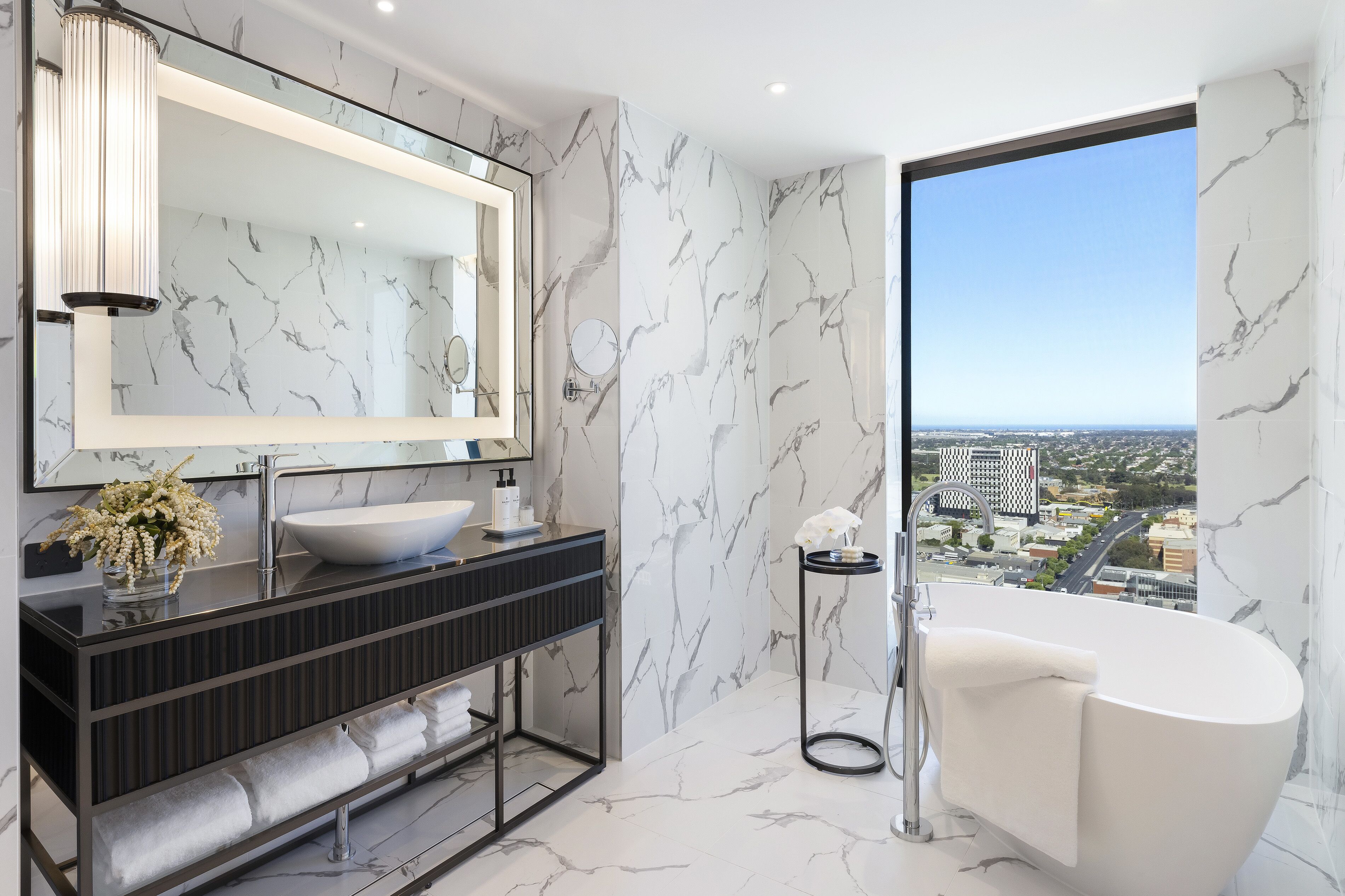 Ein modernes Badezimmer mit einem großen Spiegel, einer weißen Badewanne und Blick auf eine Stadtlandschaft durch ein großes Fenster.