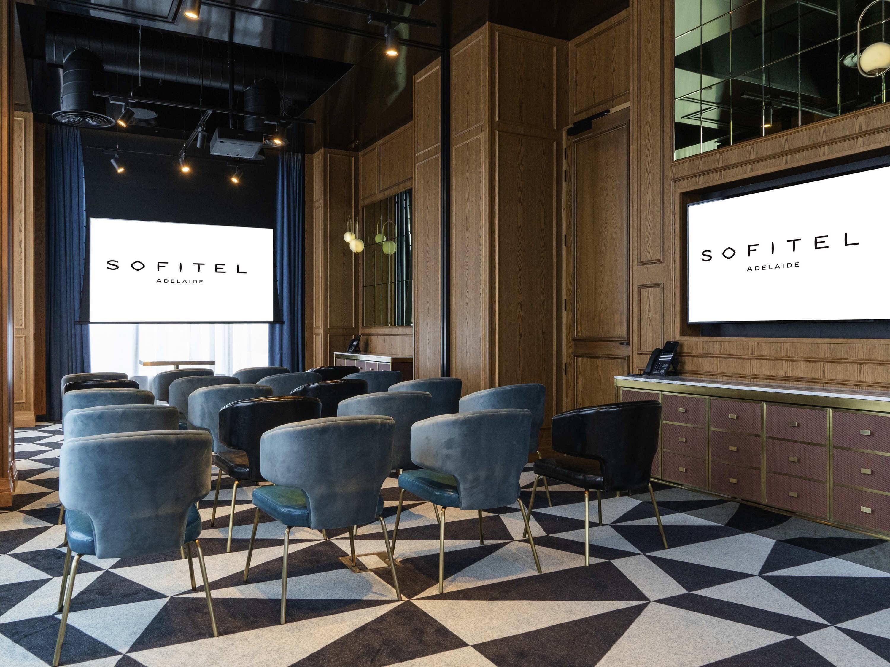 Een vergaderzaal met een vloer met geometrisch patroon, rijen stoelen en een groot scherm waarop 'SOFITEL ADELAIDE' staat.