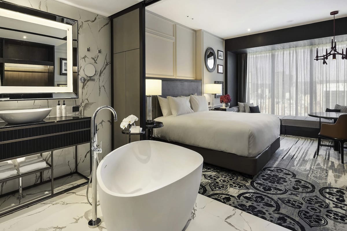 A modern hotel room with a large bed, a bathtub, a vanity with a mirror, and a dining area with a view.