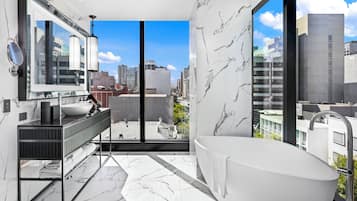 Un baño moderno con una bañera grande, un tocador con espejo y vistas a la ciudad a través de amplias ventanas.