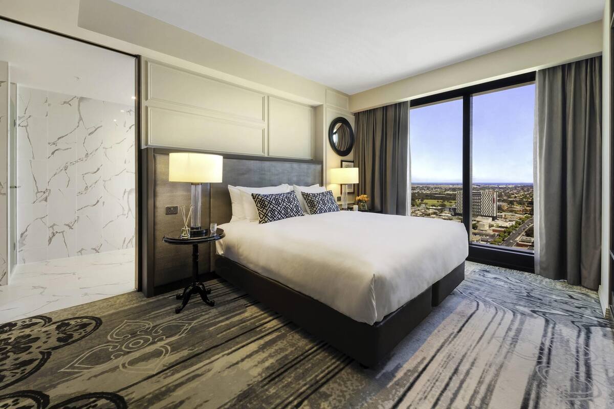 A hotel room with a large bed, a nightstand with a lamp, a mirror, and a view of the city.