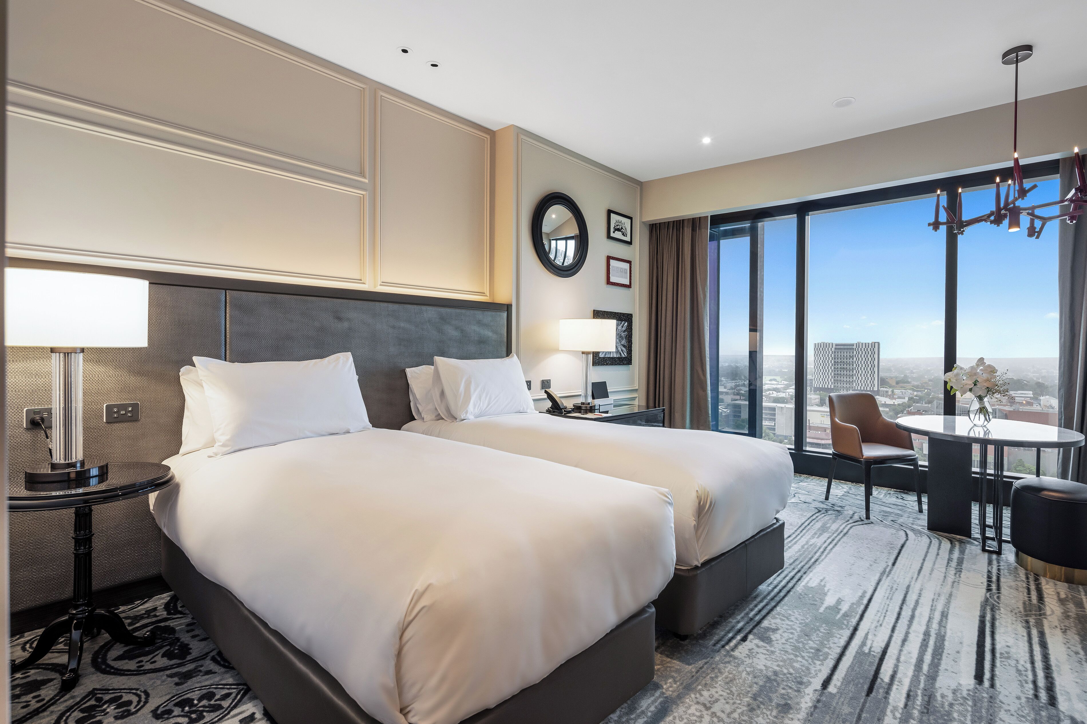 A hotel room with two beds, a desk, a chair, a round table, and a large window with a city view.