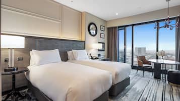 A hotel room with two beds, a desk, a chair, a round table, and a large window with a city view.