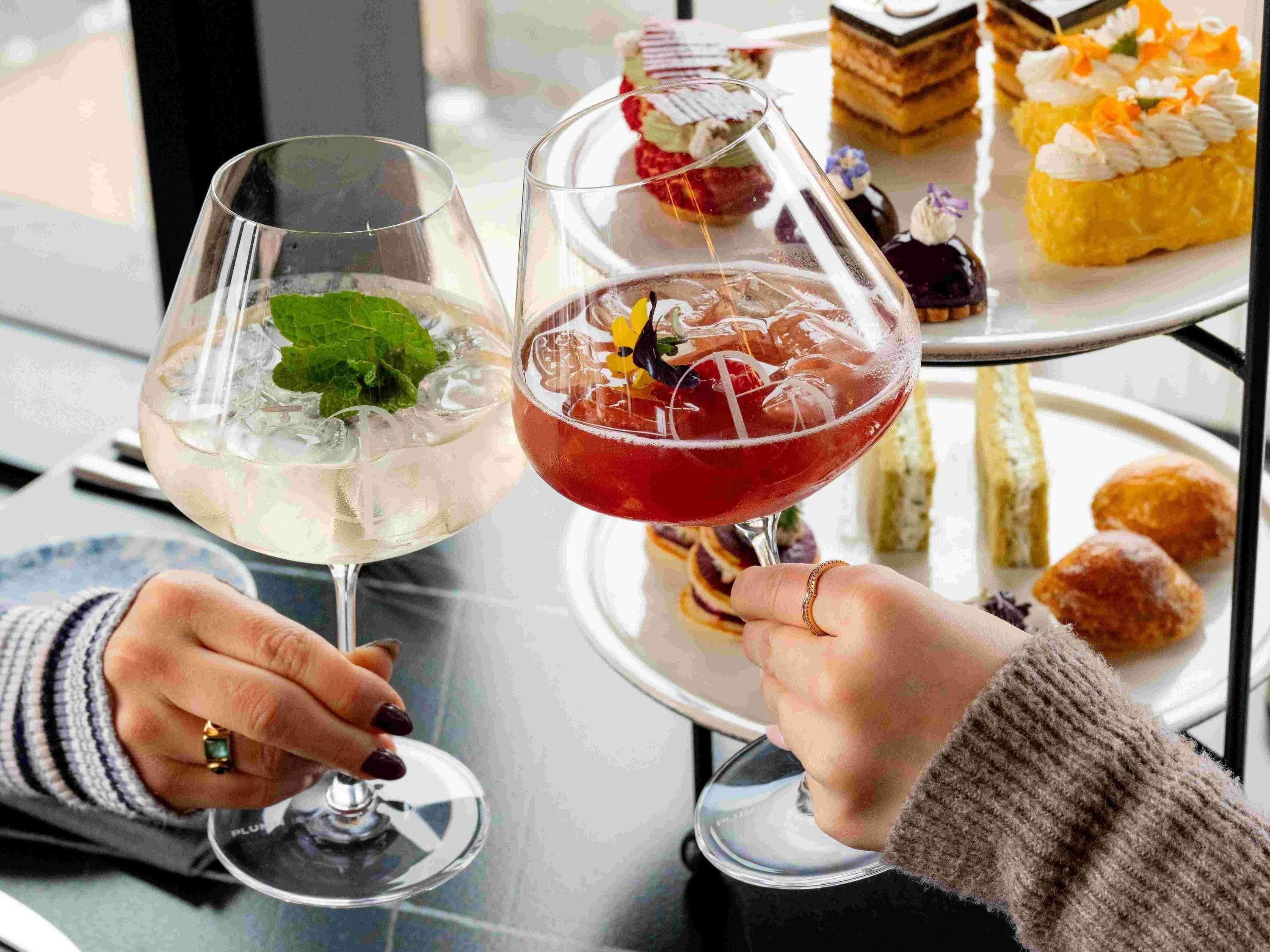 Two hands holding cocktail glasses with ice and garnishes, a dessert tray with various pastries, and a window in the background.