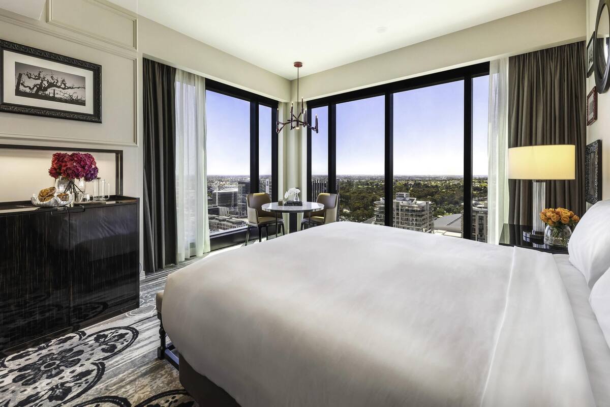 A hotel room with a large bed, a desk with a vase of flowers, a chair, and a view of the cityscape through large windows.