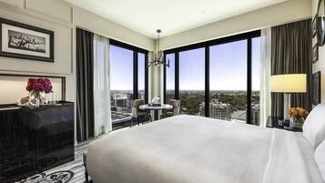 A hotel room with a large bed, a desk with a vase of flowers, a chair, and a view of the cityscape through large windows.