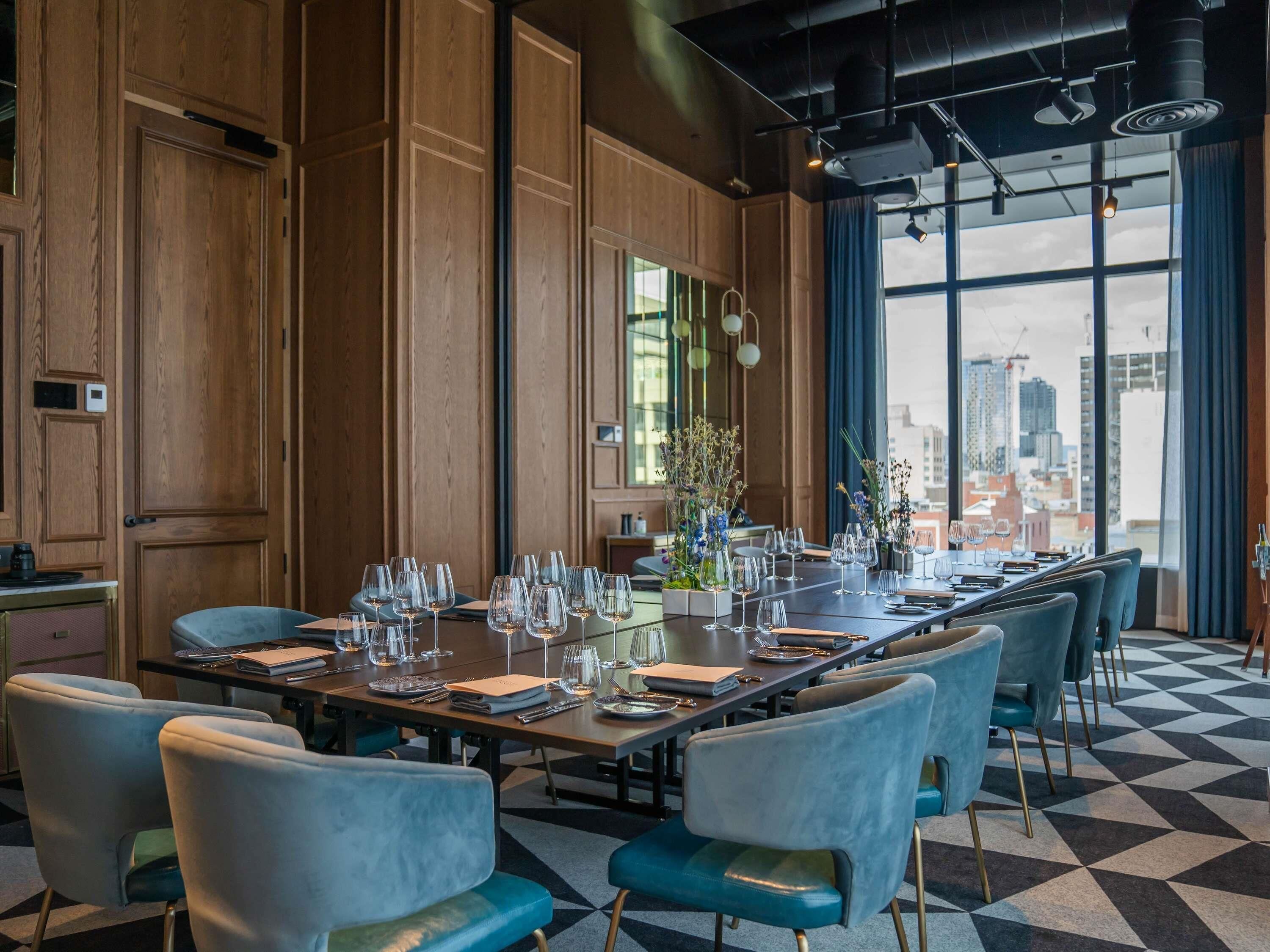 Un comedor con una larga mesa dispuesta para varios invitados, paneles de madera en las paredes y vistas de la ciudad a través de amplios ventanales.